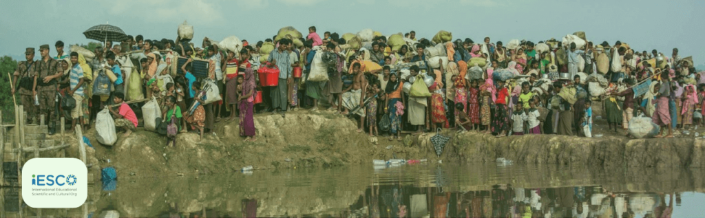 A large group of Rohingya refugees, including men, women, and children, gathered on a muddy embankment beside a body of water in Malaysia. They are carrying various belongings and are closely packed together. There are also a few soldiers or officials present, overseeing the situation. The logo of the International Educational Scientific and Cultural Organization (iESCO) is visible in the bottom left corner of the image.
