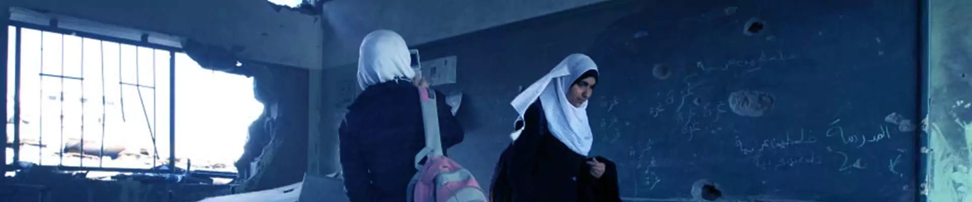 Two students in hijabs standing in a damaged classroom with broken windows and walls, illustrating the need for a Program of Emergency Intervention for Education in conflict-affected areas to restore learning environments and ensure educational continuity.