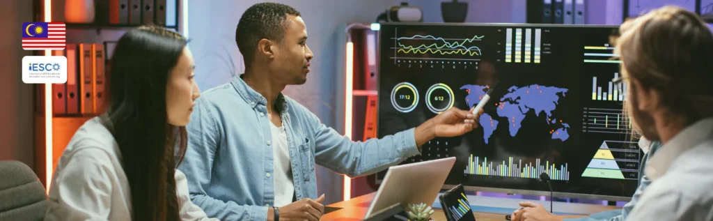 a group of three students in a modern office setting, engaged in a discussion or presentation. They are seated around a table with laptops and a small plant. One person, a man in a light blue shirt, is pointing at a large screen displaying various data visualizations.